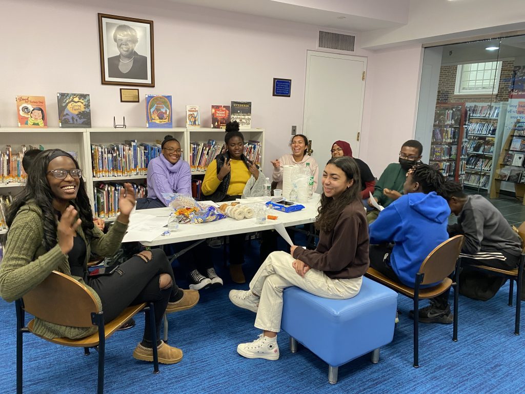 Teen volunteers gathered around a table at the Teaneck Public Library, smiling and participating in a group activity during a volunteer program meeting.